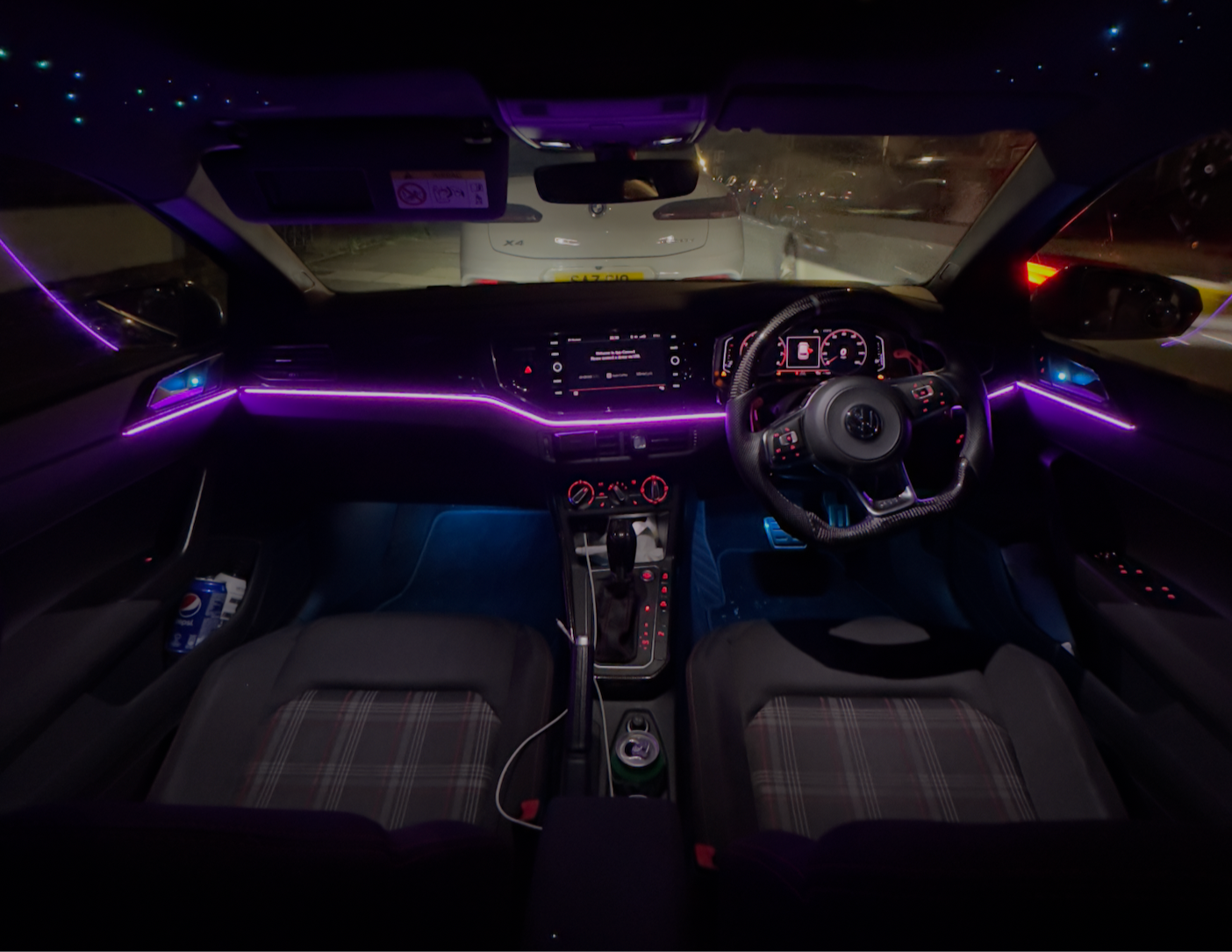 Modern car interior with purple ambient LED lighting along dashboard and doors, featuring a digital display, leather-wrapped steering wheel, checkered seats, and blue footwell lights.